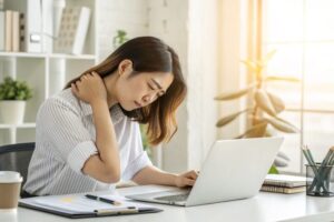 デスクワーク女性と肩こり 「PC作業で肩を押さえてつらそうにしている女性」