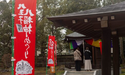 八王子七福神めぐりの赤いのぼり旗と、お堂の前で静かに手を合わせる参拝客の後ろ姿